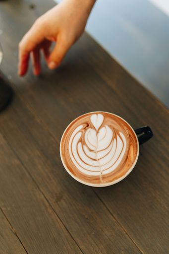 A close-up of a latte art in a cup on a wooden table with a hand reaching out.
