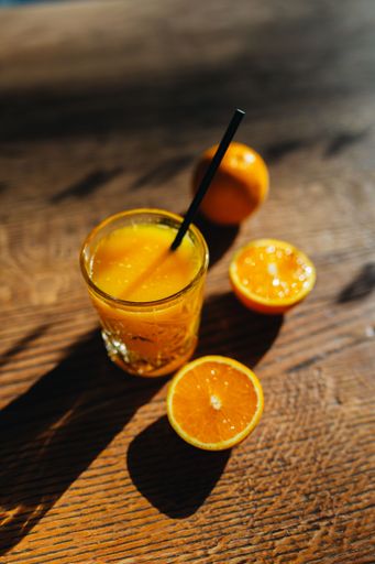 Fresh orange juice in a glass with sliced oranges on a wooden table under sunlight casting shadows.
