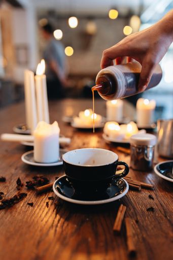 A cozy coffee shop scene with a hand pouring syrup into a black cup surrounded by candles and cinnamon sticks on a wooden table.