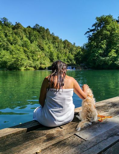 Woman and her dog sitting on edge of wooden deck by river or lake in summer. Mreznica River near Duga Resa in Croatia.