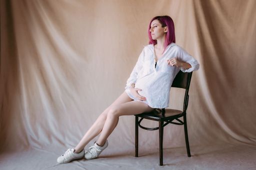 A young pregnant woman with pink hair in a white shirt sitting on a chair against a neutral backdrop