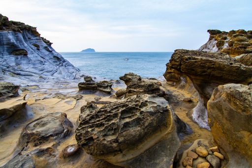 Scenic view of rocky coastline with unique geological formations and distant island under a cloudy sky.