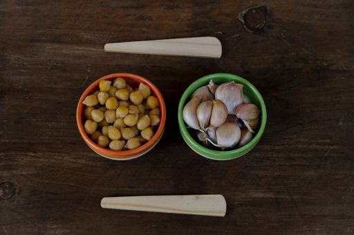 A top view shot of garlic and chickpeas in ceramic bowls with wooden spoons, hummus ingredients