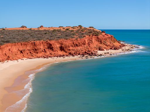 Scenic view of red cliffs meeting the turquoise ocean at a secluded beach under a clear blue sky in Western Australia.