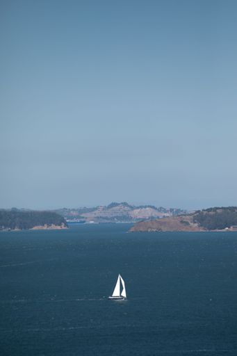 A solitary sailboat gliding across the calm waters with distant hills under a clear blue sky.