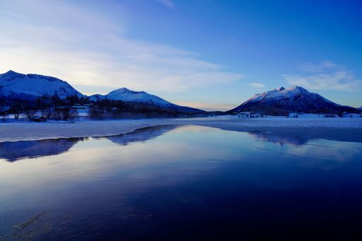 Serene winter landscape with snow-capped mountains reflecting in a calm lake under a clear blue sky.