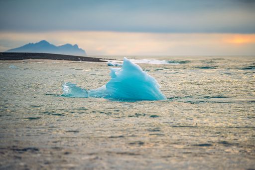 A serene view of a blue iceberg floating in calm waters with a distant mountain backdrop under a cloudy sky.