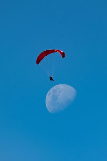 A paraglider with a red canopy soars in front of a daytime moon against a clear blue sky.