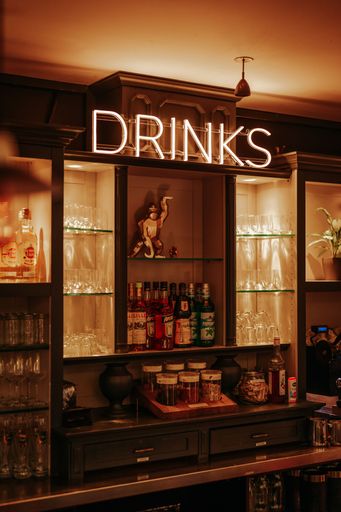 Cozy bar interior with illuminated 'Drinks' sign, showcasing a variety of spirits and glassware on shelves, creating a warm ambiance.