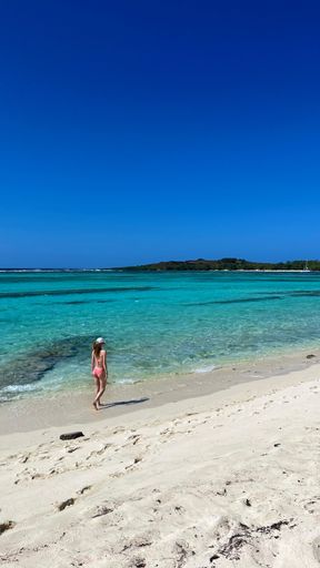 A woman in a swimsuit walking along a pristine tropical beach with clear turquoise waters under a bright blue sky.
