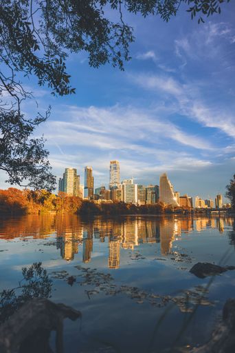 The Austin city skyline is reflected in Lady Bird Lake under a vibrant blue sky with autumn foliage framing the scene.