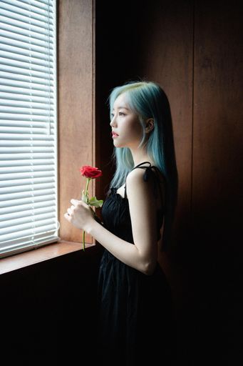 A South Korean woman with light blue hair holding a red rose and looking out the window