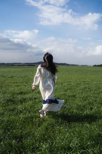 A young brunette woman wearing a light white dress and walking in a field covered in greenery