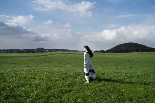 A young brunette woman wearing a light white dress and walking in a field covered in greenery
