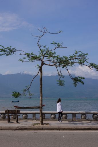 A woman in a white jacket walks along a lakeside promenade with a tree and mountains in the background under a clear blue sky.