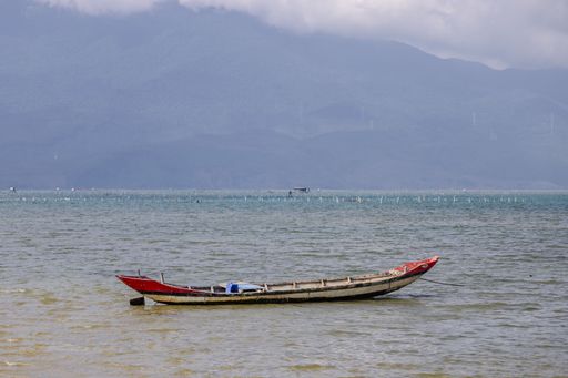 A solitary wooden boat floats on calm waters with distant mountains under a cloudy sky, creating a serene coastal scene.