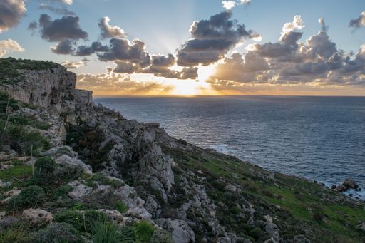 A peaceful cloudy sunset, as seen from on top a cliff along the coast in the northern part of Mellieha, Malta. A sloping cliff with boulders scree.