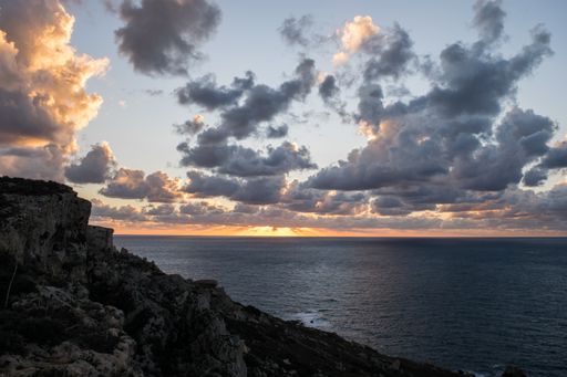 A peaceful cloudy sunset, as seen from on top a cliff along the coast in the northern part of Mellieha, Malta. A sloping cliff with boulders scree.