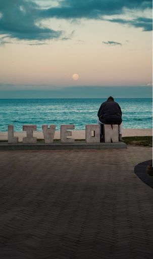 A solitary figure sits by the ocean beside 'Live On' letters at sunset, reflecting tranquility and introspection.