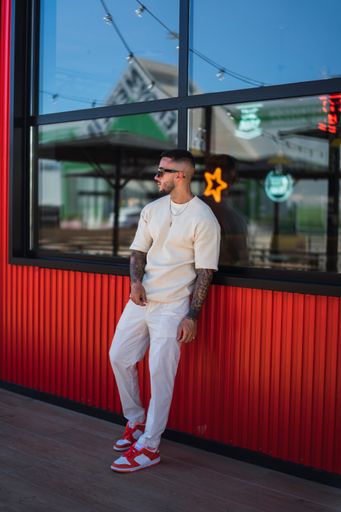 A stylish guy with sunglasses and tattoos leaning on the building with a reflective window