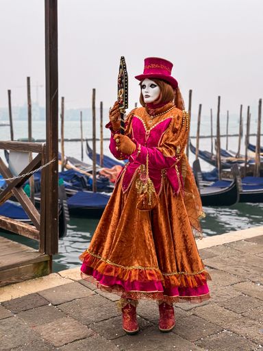 Person in vibrant Venetian carnival costume with mask and mirror by gondolas, capturing the essence of Venice's cultural festival.