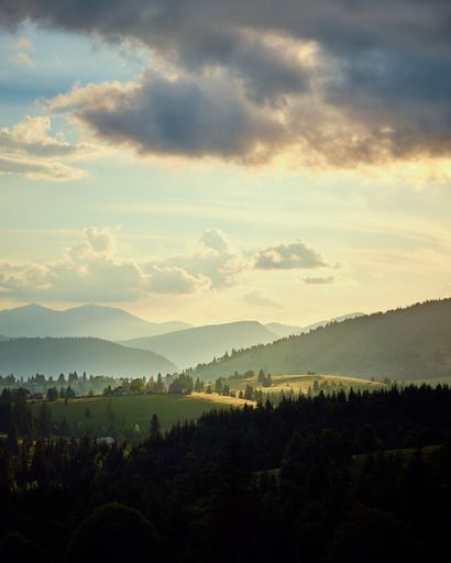 A vertical shot of hills covered in forests under the sunlight and a cloudy sky in the countryside