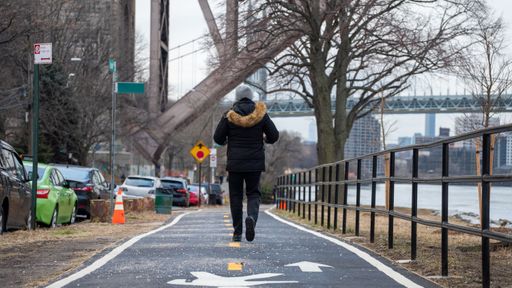 A person in a winter coat walks along a riverside path in an urban setting with a bridge in the background.