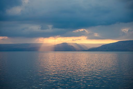 The sunlight breaks through clouds over a tranquil lake with distant hills at sunset, creating a serene and picturesque scene.