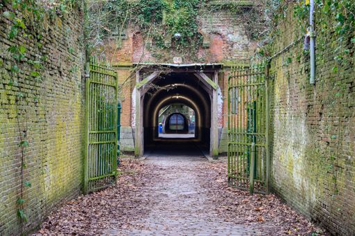 An old brick tunnel entrance with open iron gates, surrounded by moss-covered walls and fallen leaves, creating a mysterious atmosphere.