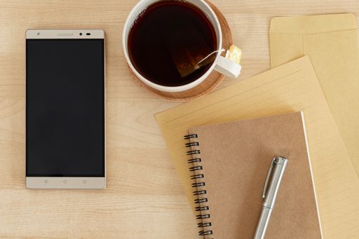 A top view of a tablet, notebook, recycled paper, pen, and teacup on a wooden desk