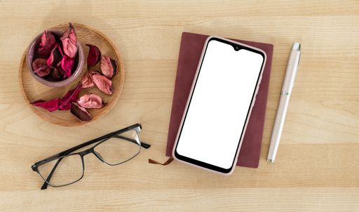 A top view of blank mobile phone, notebook, pen, eyeglasses on a wooden office desk