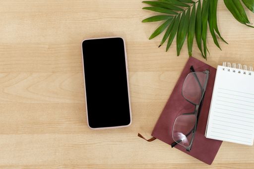 A top view of a blank smartphone, notebook, and eyeglasses on a wooden office desk