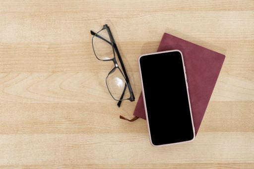 A top view of a blank smartphone, notebook, and eyeglasses on a wooden office desk