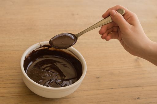 A shot of a hand taking melted chocolate with a spoon from a white ceramic bowl on a wooden table
