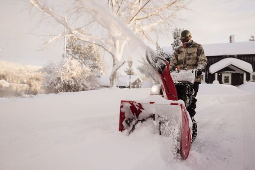 A man in winter gear operates a snowblower on a snowy driveway in a rural setting. Norway