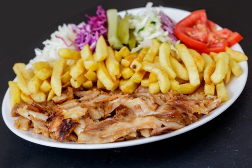 A closeup of Chiken Doner Kebab on a plate with french fries, tomatoes, onion, pickles and cabbage on a black surface