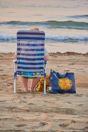 A vertical shot of an old woman sitting on a folding chair by the sea and enjoying the view