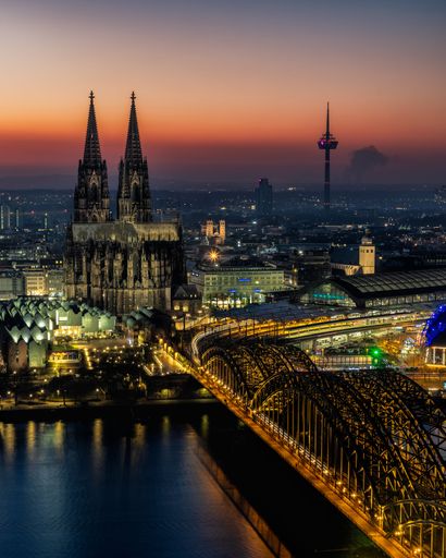 Stunning view of Cologne Cathedral and Hohenzollern Bridge at sunset, with city lights reflecting on the Rhine River in Germany.