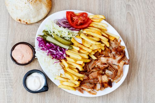 A top view of Chicken Doner Kebab on a plate with bread, sauce, french fries, tomatoes, onion, pickles and cabbage on a wooden table