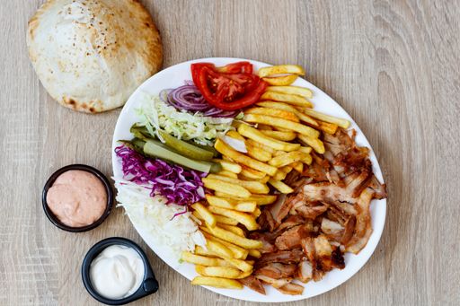 A top view of Chicken Doner Kebab on a plate with bread, sauce, french fries, tomatoes, onion, pickles and cabbage on a wooden table