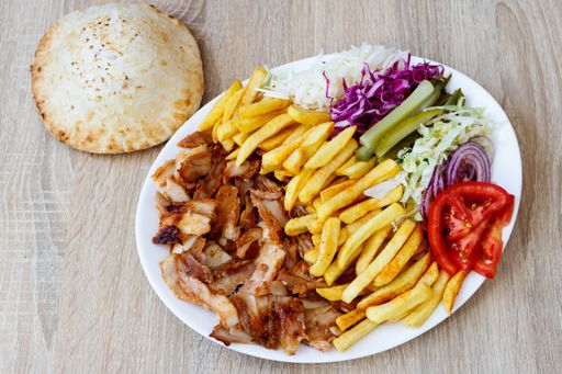 A top view of Chicken Doner Kebab on a plate with bread, french fries, tomatoes, onion, pickles and cabbage on a wooden table