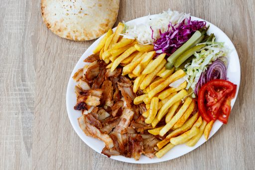 A top view of Chiken Doner Kebab on a plate with bread, french fries, tomatoes, onion, pickles and cabbage on a wooden table