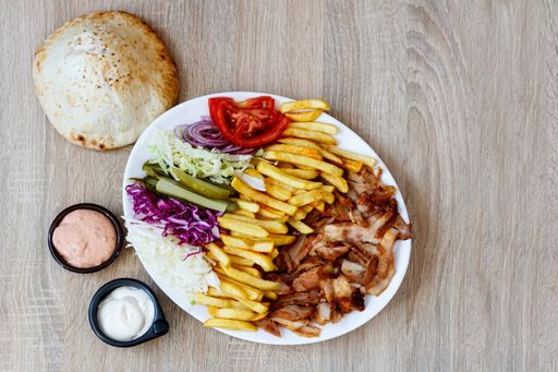 A top view of Chiken Doner Kebab on a plate with bread, sauce, french fries, tomatoes, onion, pickles and cabbage on a wooden table