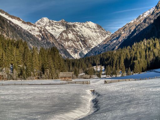 A serene winter landscape showcasing snow-covered fields, a rustic wooden barn, and towering mountains under a clear blue sky.