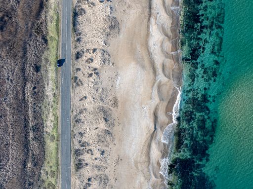 An aerial view of a scenic coastal road alongside a sandy beach with clear blue waters under a bright sky