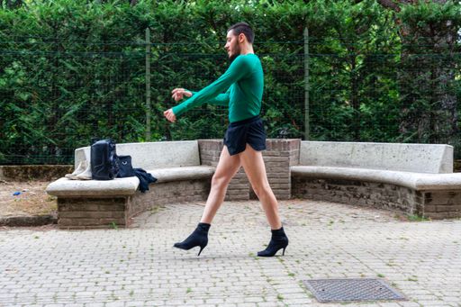 A young Caucasian man wearing high heels, shorts and a green shirt while posing in a park