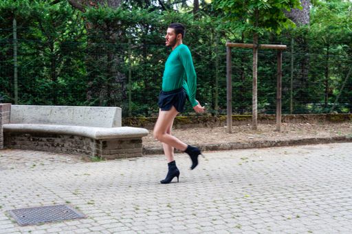 A young Caucasian man wearing high heels, shorts and a green shirt while posing in a park