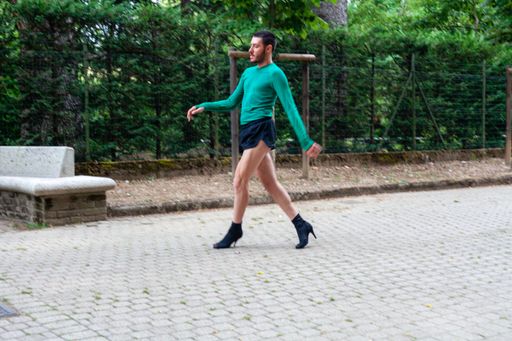 A young Caucasian man wearing high heels, shorts and a green shirt while posing in a park