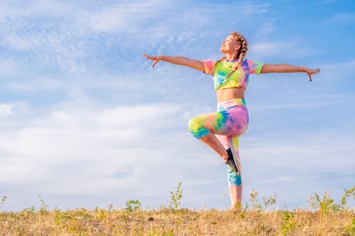Beautiful girl gymnast in a bright tracksuit goes in for sports outdoors. Stands on one leg, arms wide apart, looks forward in front of him.