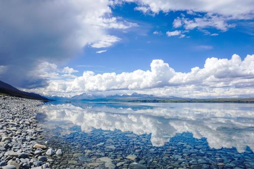 A serene lake with a rocky shore reflecting fluffy clouds and distant mountains under a blue sky.